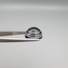 Close-up photograph of a 6.28-carat natural black eye agate cabochon gemstone held in stainless steel tweezers against a neutral grey background, featuring crisp white concentric eye rings framing a deep black center and a translucent back base.