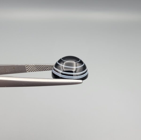 Close-up photograph of a 6.28-carat natural black eye agate cabochon gemstone held in stainless steel tweezers against a neutral grey background, featuring crisp white concentric eye rings framing a deep black center and a translucent back base.