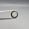 Close-up photograph of a 6.28-carat natural black eye agate cabochon gemstone held in stainless steel tweezers against a neutral grey background, featuring crisp white concentric eye rings framing a deep black center and a translucent back base.