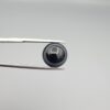 Close-up photograph of a 6.55-carat natural black eye agate cabochon gemstone held in stainless steel tweezers against a neutral grey background, featuring crisp white concentric eye rings framing a deep black center.
