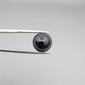 Close-up photograph of a 6.55-carat natural black eye agate cabochon gemstone held in stainless steel tweezers against a neutral grey background, featuring crisp white concentric eye rings framing a deep black center.