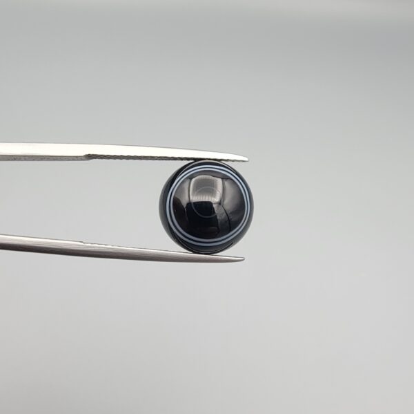 Close-up photograph of a 6.55-carat natural black eye agate cabochon gemstone held in stainless steel tweezers against a neutral grey background, featuring crisp white concentric eye rings framing a deep black center.