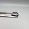 Close-up photograph of a 6.55-carat natural black eye agate cabochon gemstone held in stainless steel tweezers against a neutral grey background, featuring crisp white concentric eye rings framing a deep black center.