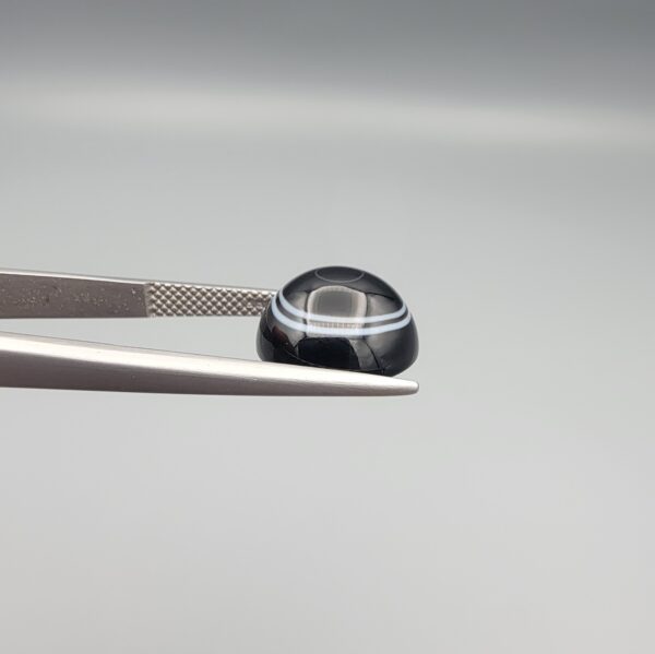 Close-up photograph of a 6.55-carat natural black eye agate cabochon gemstone held in stainless steel tweezers against a neutral grey background, featuring crisp white concentric eye rings framing a deep black center.