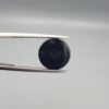 Close-up photograph of a 6.55-carat natural black eye agate cabochon gemstone held in stainless steel tweezers against a neutral grey background, featuring crisp white concentric eye rings framing a deep black center.