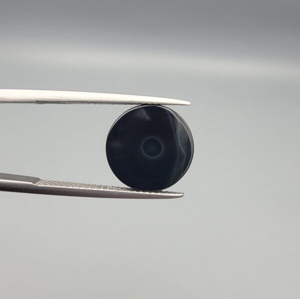 Close-up photograph of a 6.55-carat natural black eye agate cabochon gemstone held in stainless steel tweezers against a neutral grey background, featuring crisp white concentric eye rings framing a deep black center.