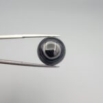 Close-up photograph of a 12.75-carat natural black eye agate cabochon gemstone held in stainless steel tweezers against a neutral grey background, showcasing a prominent concentric white eye pattern set against an intense black center and a patterned translucent greyish-blue reverse.