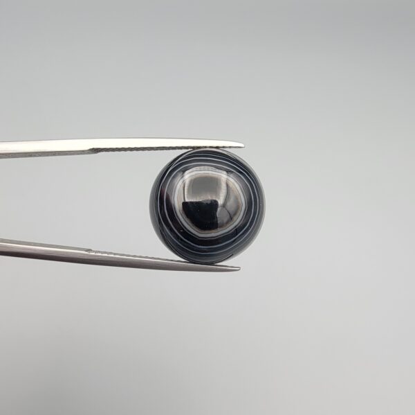 Close-up photograph of a 12.75-carat natural black eye agate cabochon gemstone held in stainless steel tweezers against a neutral grey background, showcasing a prominent concentric white eye pattern set against an intense black center and a patterned translucent greyish-blue reverse.