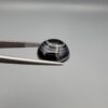 Close-up photograph of a 12.75-carat natural black eye agate cabochon gemstone held in stainless steel tweezers against a neutral grey background, showcasing a prominent concentric white eye pattern set against an intense black center and a patterned translucent greyish-blue reverse.