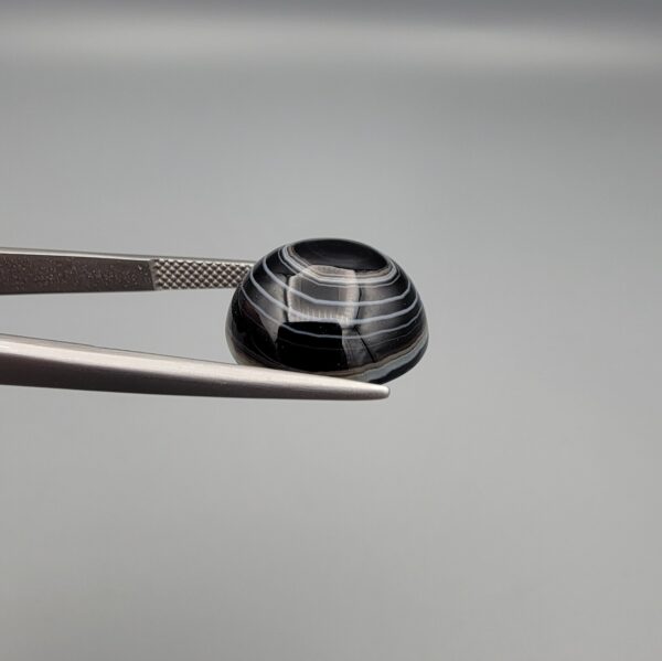 Close-up photograph of a 12.75-carat natural black eye agate cabochon gemstone held in stainless steel tweezers against a neutral grey background, showcasing a prominent concentric white eye pattern set against an intense black center and a patterned translucent greyish-blue reverse.
