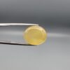 Close-up photograph of an 11.95-carat natural yellow chalcedony agate gemstone held in stainless steel tweezers against a neutral grey background, showcasing a translucent oval cabochon shape with a warm golden hue and high-polish finish.
