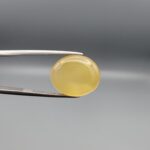 Close-up photograph of an 11.95-carat natural yellow chalcedony agate gemstone held in stainless steel tweezers against a neutral grey background, showcasing a translucent oval cabochon shape with a warm golden hue and high-polish finish.