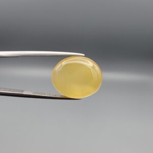 Close-up photograph of an 11.95-carat natural yellow chalcedony agate gemstone held in stainless steel tweezers against a neutral grey background, showcasing a translucent oval cabochon shape with a warm golden hue and high-polish finish.