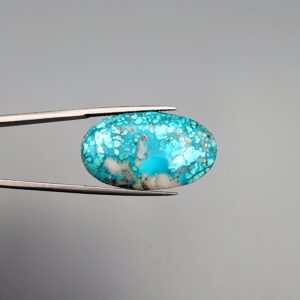 Close-up photograph of a 36.88-carat natural sky-blue Persian turquoise oval cabochon held in stainless steel tweezers against a neutral grey background, featuring vibrant blue tones, striking white host-rock patches, and glittering metallic pyrite inclusions.