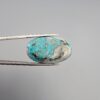 Close-up photograph of a 36.88-carat natural sky-blue Persian turquoise oval cabochon held in stainless steel tweezers against a neutral grey background, featuring vibrant blue tones, striking white host-rock patches, and glittering metallic pyrite inclusions.