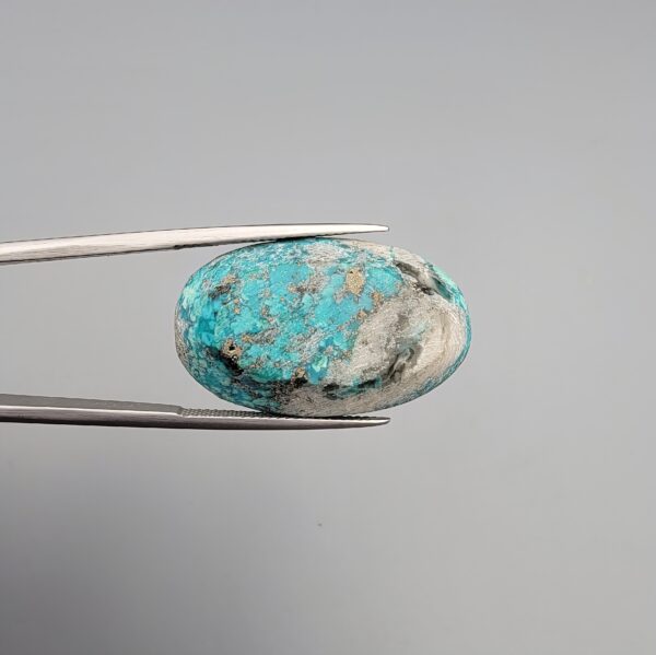 Close-up photograph of a 36.88-carat natural sky-blue Persian turquoise oval cabochon held in stainless steel tweezers against a neutral grey background, featuring vibrant blue tones, striking white host-rock patches, and glittering metallic pyrite inclusions.