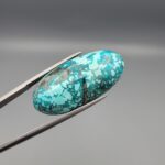 Close-up photograph of a 32.46-carat natural sky-blue Persian turquoise gemstone resting on a human hand, showcasing an elongated oval cabochon cut with a webbed dark matrix and glittering pyrite inclusions.