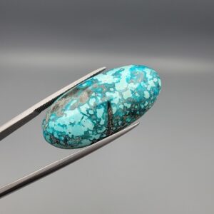 Close-up photograph of a 32.46-carat natural sky-blue Persian turquoise gemstone resting on a human hand, showcasing an elongated oval cabochon cut with a webbed dark matrix and glittering pyrite inclusions.