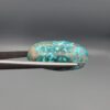 Close-up photograph of a 42.30-carat natural deep blue mottled Persian turquoise oval cabochon held in stainless steel tweezers against a neutral background, highlighting its brownish-grey matrix, metallic pyrite inclusions, and natural host-rock backing.