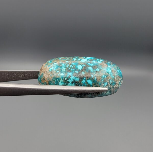 Close-up photograph of a 42.30-carat natural deep blue mottled Persian turquoise oval cabochon held in stainless steel tweezers against a neutral background, highlighting its brownish-grey matrix, metallic pyrite inclusions, and natural host-rock backing.