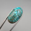 Close-up photograph of a 42.30-carat natural deep blue mottled Persian turquoise oval cabochon held in stainless steel tweezers against a neutral background, highlighting its brownish-grey matrix, metallic pyrite inclusions, and natural host-rock backing.