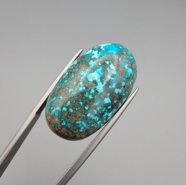 Close-up photograph of a 42.30-carat natural deep blue mottled Persian turquoise oval cabochon held in stainless steel tweezers against a neutral background, highlighting its brownish-grey matrix, metallic pyrite inclusions, and natural host-rock backing.