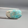 Close-up photograph of a 42.30-carat natural deep blue mottled Persian turquoise oval cabochon held in stainless steel tweezers against a neutral background, highlighting its brownish-grey matrix, metallic pyrite inclusions, and natural host-rock backing.