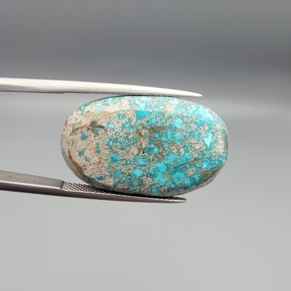 Close-up photograph of a 42.30-carat natural deep blue mottled Persian turquoise oval cabochon held in stainless steel tweezers against a neutral background, highlighting its brownish-grey matrix, metallic pyrite inclusions, and natural host-rock backing.