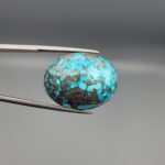Close-up photograph of a 45.95-carat natural robin's-egg blue Persian turquoise oval cabochon held in stainless steel tweezers against a neutral background, highlighting its vibrant blue tone, heavy metallic pyrite patches, and organic matrix backing.