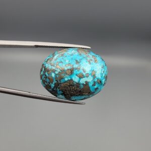 Close-up photograph of a 45.95-carat natural robin's-egg blue Persian turquoise oval cabochon held in stainless steel tweezers against a neutral background, highlighting its vibrant blue tone, heavy metallic pyrite patches, and organic matrix backing.
