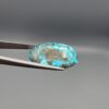 Close-up photograph of a 45.95-carat natural robin's-egg blue Persian turquoise oval cabochon held in stainless steel tweezers against a neutral background, highlighting its vibrant blue tone, heavy metallic pyrite patches, and organic matrix backing.