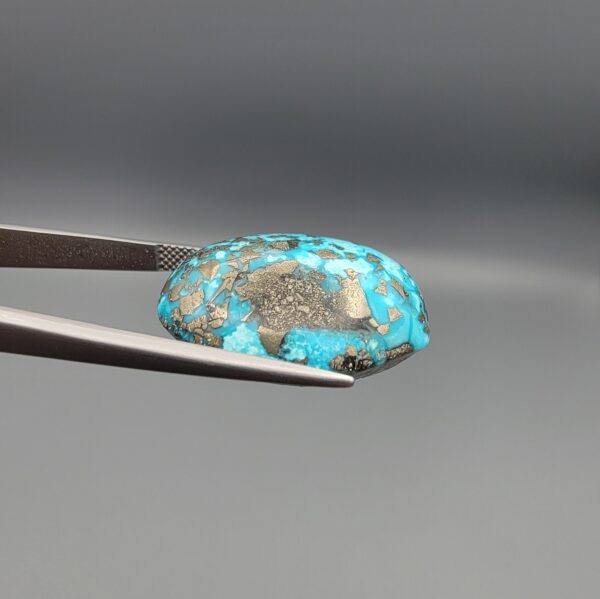 Close-up photograph of a 45.95-carat natural robin's-egg blue Persian turquoise oval cabochon held in stainless steel tweezers against a neutral background, highlighting its vibrant blue tone, heavy metallic pyrite patches, and organic matrix backing.