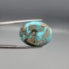 Close-up photograph of a 45.95-carat natural robin's-egg blue Persian turquoise oval cabochon held in stainless steel tweezers against a neutral background, highlighting its vibrant blue tone, heavy metallic pyrite patches, and organic matrix backing.
