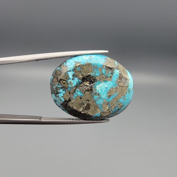 Close-up photograph of a 45.95-carat natural robin's-egg blue Persian turquoise oval cabochon held in stainless steel tweezers against a neutral background, highlighting its vibrant blue tone, heavy metallic pyrite patches, and organic matrix backing.