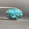 ​Close-up photograph of a 32.56-carat natural robin's-egg blue Persian turquoise oval cabochon held in stainless steel tweezers against a neutral background, highlighting its vibrant blue hue and shimmering scattered metallic pyrite patches.