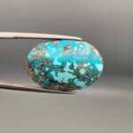 ​Close-up photograph of a 32.56-carat natural robin's-egg blue Persian turquoise oval cabochon held in stainless steel tweezers against a neutral background, highlighting its vibrant blue hue and shimmering scattered metallic pyrite patches.