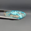 ​Close-up photograph of a 32.56-carat natural robin's-egg blue Persian turquoise oval cabochon held in stainless steel tweezers against a neutral background, highlighting its vibrant blue hue and shimmering scattered metallic pyrite patches.