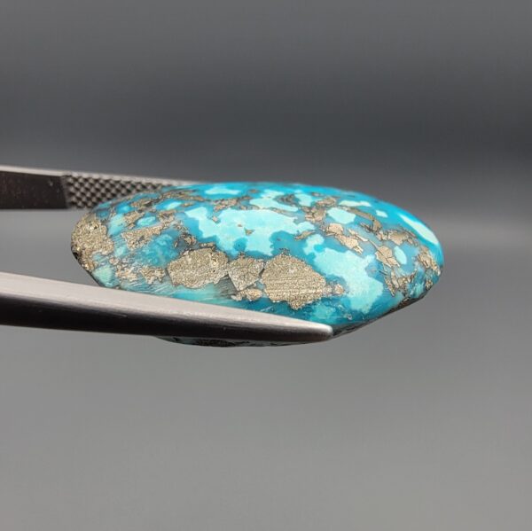 ​Close-up photograph of a 32.56-carat natural robin's-egg blue Persian turquoise oval cabochon held in stainless steel tweezers against a neutral background, highlighting its vibrant blue hue and shimmering scattered metallic pyrite patches.