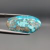 ​Close-up photograph of a 32.56-carat natural robin's-egg blue Persian turquoise oval cabochon held in stainless steel tweezers against a neutral background, highlighting its vibrant blue hue and shimmering scattered metallic pyrite patches.