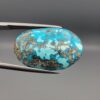 ​Close-up photograph of a 32.56-carat natural robin's-egg blue Persian turquoise oval cabochon held in stainless steel tweezers against a neutral background, highlighting its vibrant blue hue and shimmering scattered metallic pyrite patches.