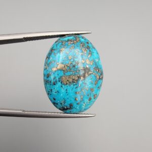 Close-up photograph of a 24.95-carat natural sky-blue Persian turquoise oval cabochon held in stainless steel tweezers against a neutral grey background, showcasing vibrant sky-blue tones, abundant metallic pyrite speckles, and dark matrix inclusions.