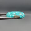 Close-up photograph of a 44.26-carat natural bright sky-blue Persian turquoise oval cabochon held in stainless steel tweezers against a neutral grey background, showcasing vibrant sky-blue saturation and distinct metallic pyrite patches.