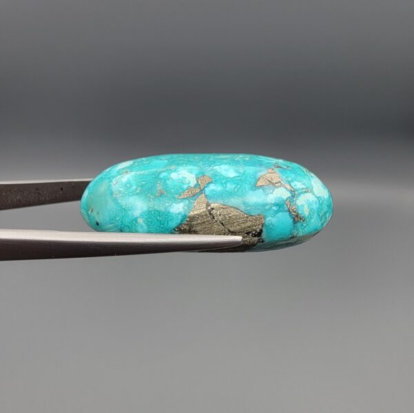 Close-up photograph of a 44.26-carat natural bright sky-blue Persian turquoise oval cabochon held in stainless steel tweezers against a neutral grey background, showcasing vibrant sky-blue saturation and distinct metallic pyrite patches.