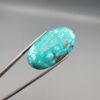 Close-up photograph of a 44.26-carat natural bright sky-blue Persian turquoise oval cabochon held in stainless steel tweezers against a neutral grey background, showcasing vibrant sky-blue saturation and distinct metallic pyrite patches.