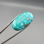 Close-up photograph of a 44.26-carat natural bright sky-blue Persian turquoise oval cabochon held in stainless steel tweezers against a neutral grey background, showcasing vibrant sky-blue saturation and distinct metallic pyrite patches.