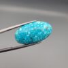 Close-up photograph of a 19.16-carat natural bright sky-blue Persian turquoise oval cabochon held in stainless steel tweezers against a neutral grey background, showcasing vibrant sky-blue color and subtle metallic pyrite flecks.
