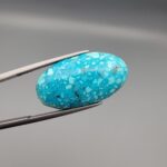 Close-up photograph of a 19.16-carat natural bright sky-blue Persian turquoise oval cabochon held in stainless steel tweezers against a neutral grey background, showcasing vibrant sky-blue color and subtle metallic pyrite flecks.