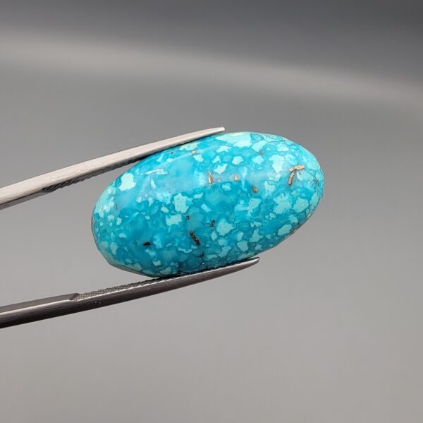 Close-up photograph of a 19.16-carat natural bright sky-blue Persian turquoise oval cabochon held in stainless steel tweezers against a neutral grey background, showcasing vibrant sky-blue color and subtle metallic pyrite flecks.