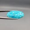 Close-up photograph of a 19.16-carat natural bright sky-blue Persian turquoise oval cabochon held in stainless steel tweezers against a neutral grey background, showcasing vibrant sky-blue color and subtle metallic pyrite flecks.