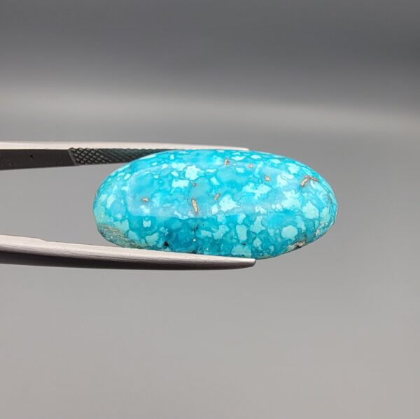 Close-up photograph of a 19.16-carat natural bright sky-blue Persian turquoise oval cabochon held in stainless steel tweezers against a neutral grey background, showcasing vibrant sky-blue color and subtle metallic pyrite flecks.